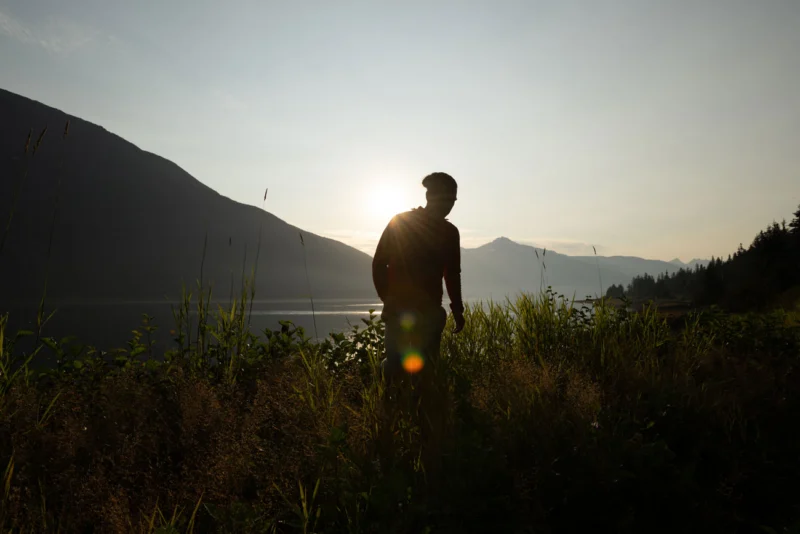 A person stands silhouetted in tall grass, backlit by the sun over a lake with mountains in the background.