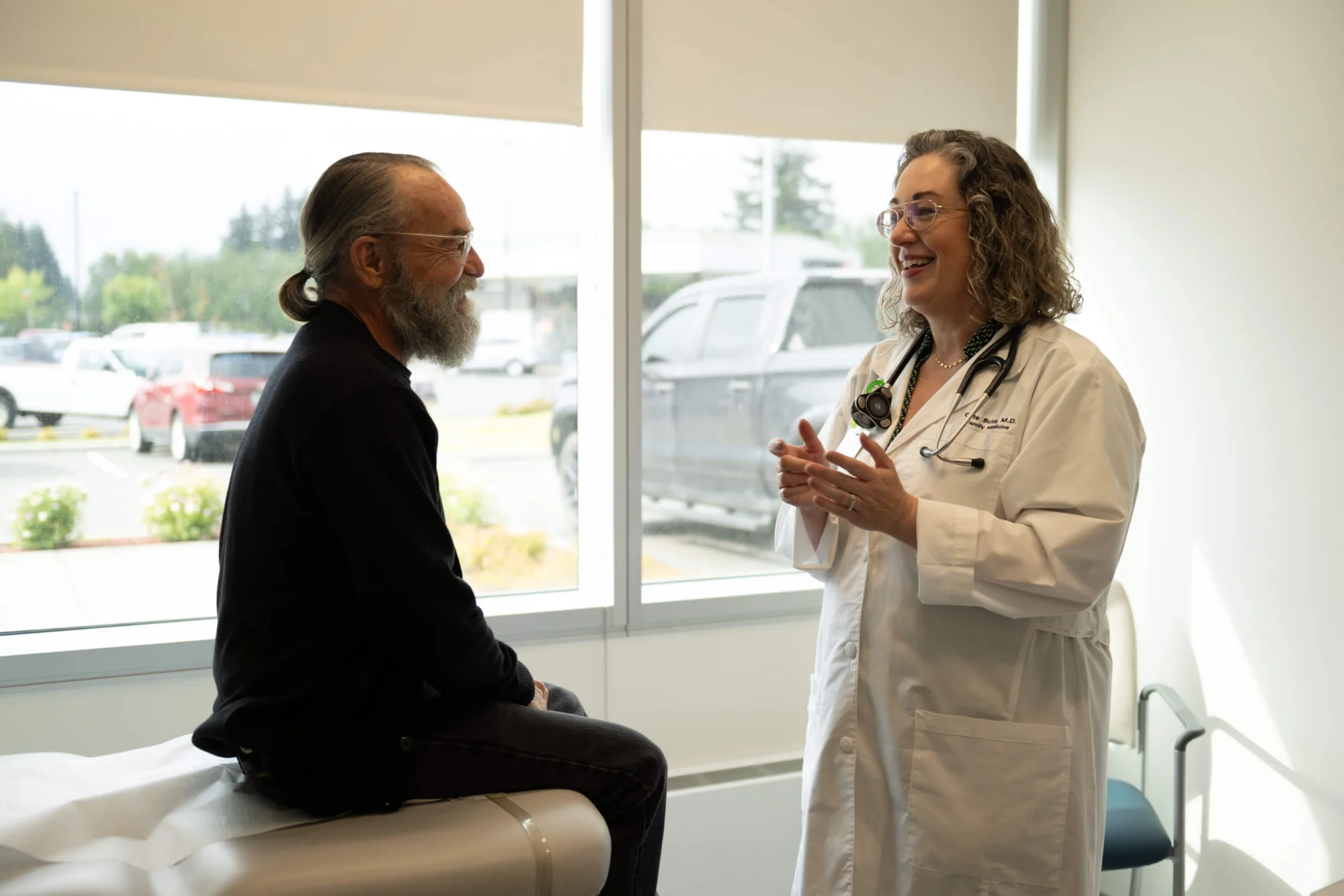 A smiling female doctor in a white lab coat talks to a smiling male patient sitting on an examination table in a bright room with windows.