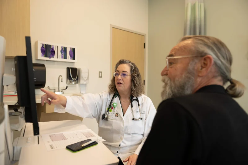 A female doctor in a white lab coat points at a computer monitor while speaking to a male patient with a beard and ponytail in a medical office.