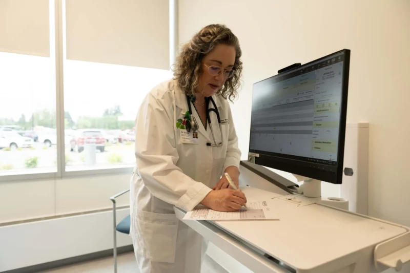 A female doctor in a white lab coat writing on a form while looking at a computer screen in a medical office.
