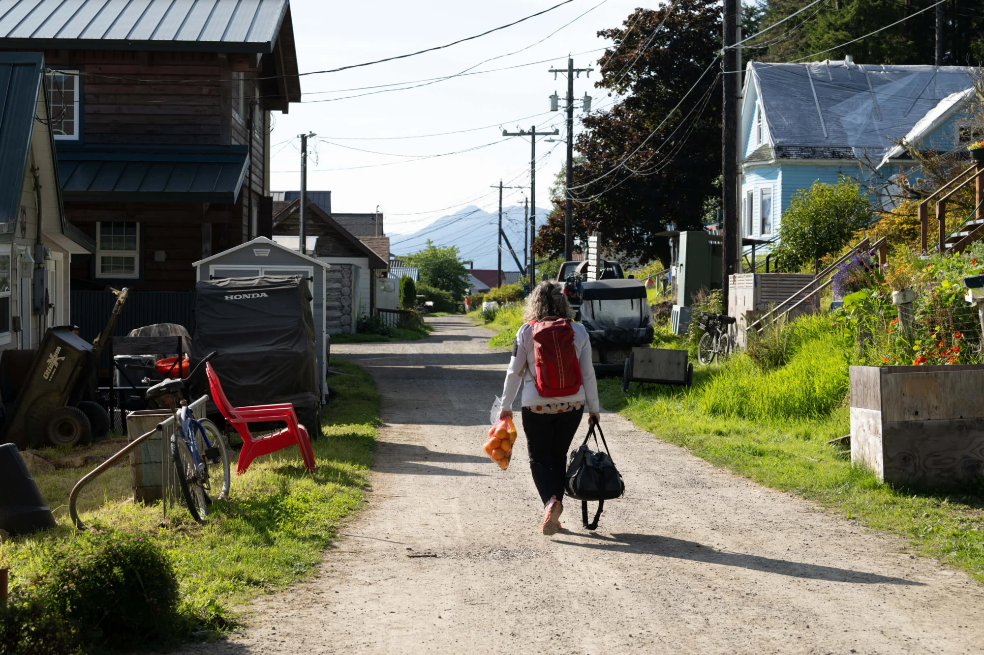 Rear view of a person with a red backpack walking down a dirt path, carrying a bag of oranges and a duffel bag. Houses, green grass, and distant mountains are visible under a bright sky.