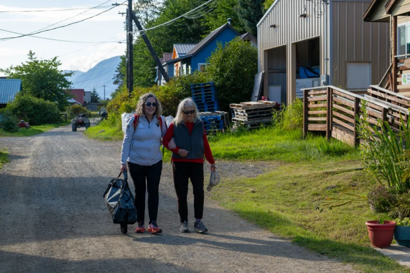 Two women, one younger with curly hair and one older with white hair, walk arm-in-arm down a gravel road. They are both wearing sunglasses and casual attire. Houses, trees, and mountains are visible in the background.