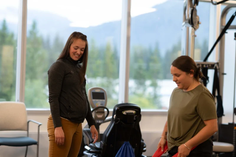 Two women in a physical therapy gym. One woman in a grey jacket and yellow pants smiles while looking at another woman in an olive top holding a red resistance band. Mountains are visible through large windows in the background.
