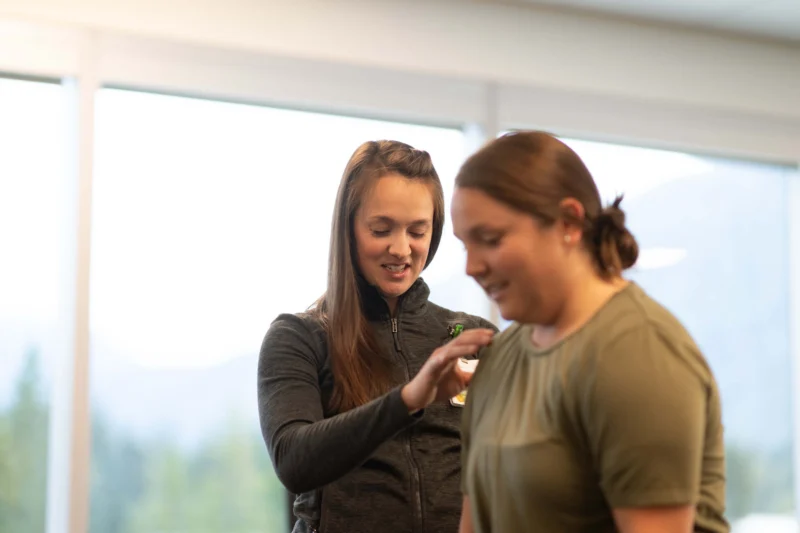 Two women, one in a gray jacket with long brown hair, places her hand on the shoulder of another woman in an olive t-shirt, both smiling indoors with a blurred outdoor background.