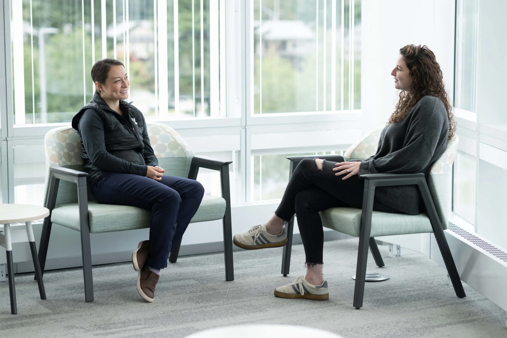 Two women sitting in modern chairs, facing each other and smiling, in a room with large windows overlooking green trees.