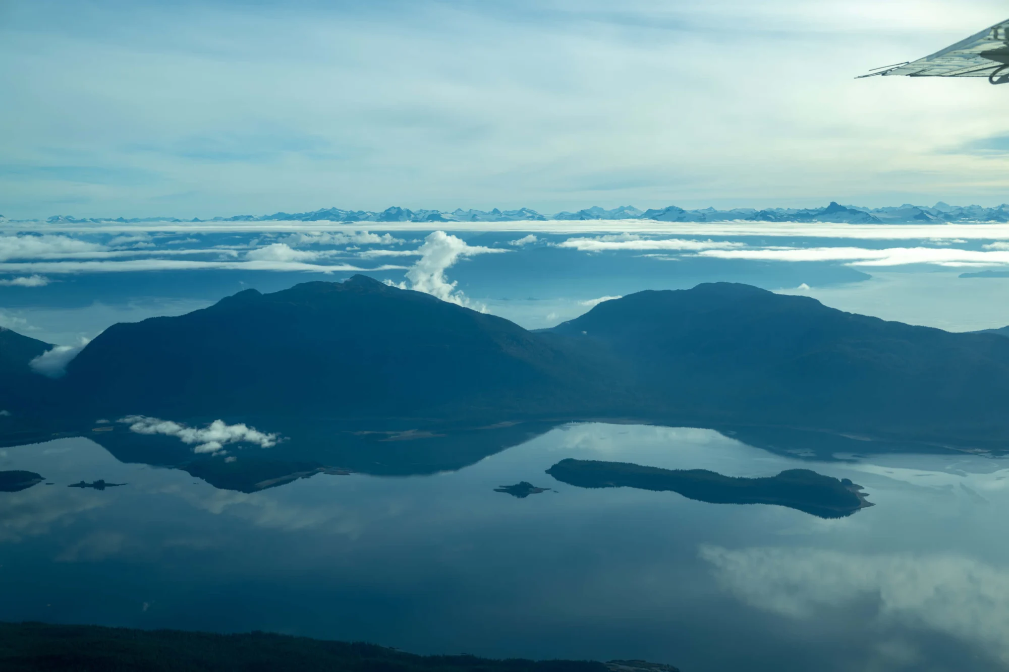 Aerial view from an airplane window showing dark mountains reflected in calm blue water, with distant snow-capped mountains and clouds on the horizon. A part of an airplane wing is visible in the upper right.