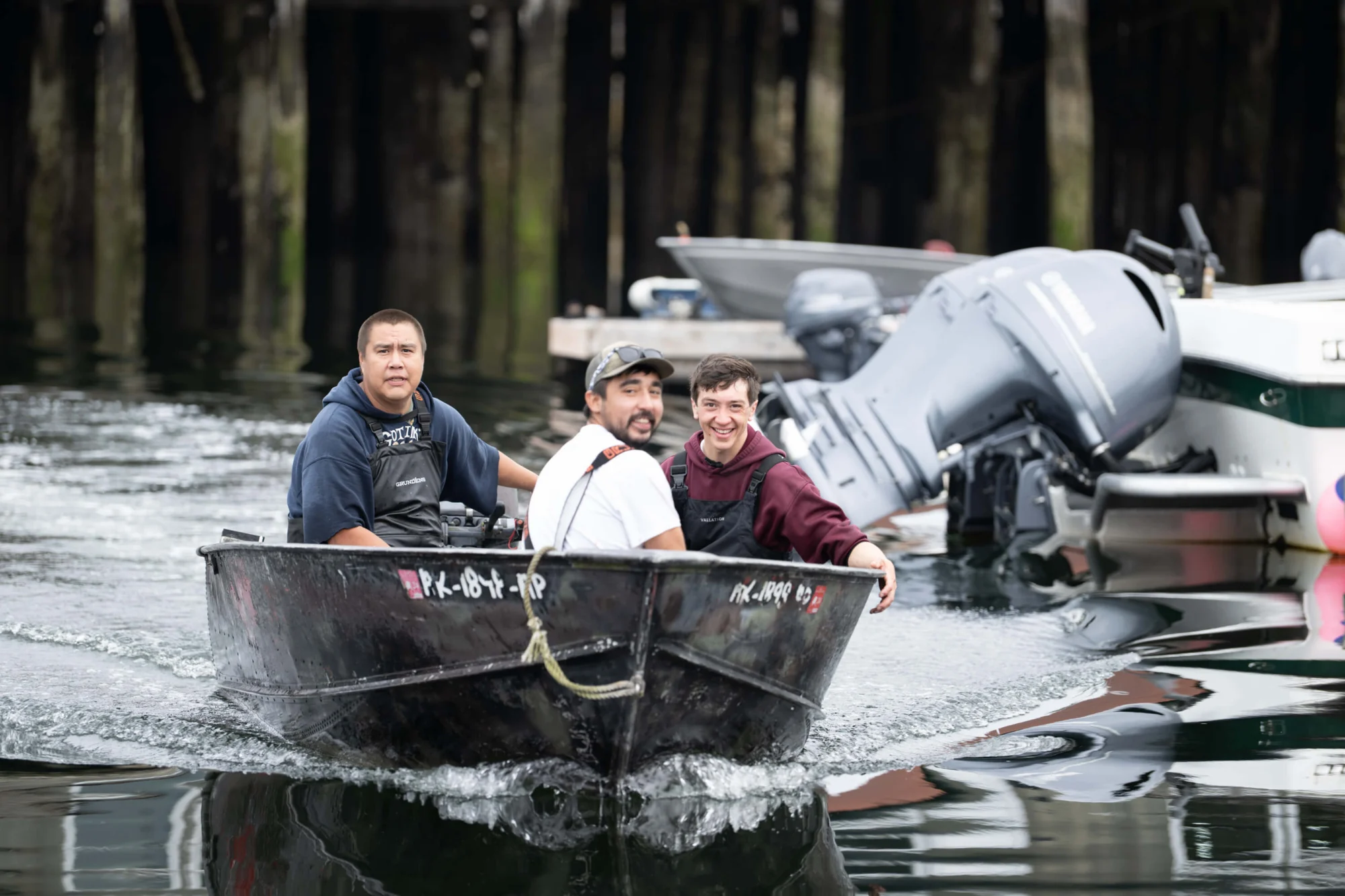 Three men in a small boat on the water, smiling. Dock pilings and other boats are in the background.
