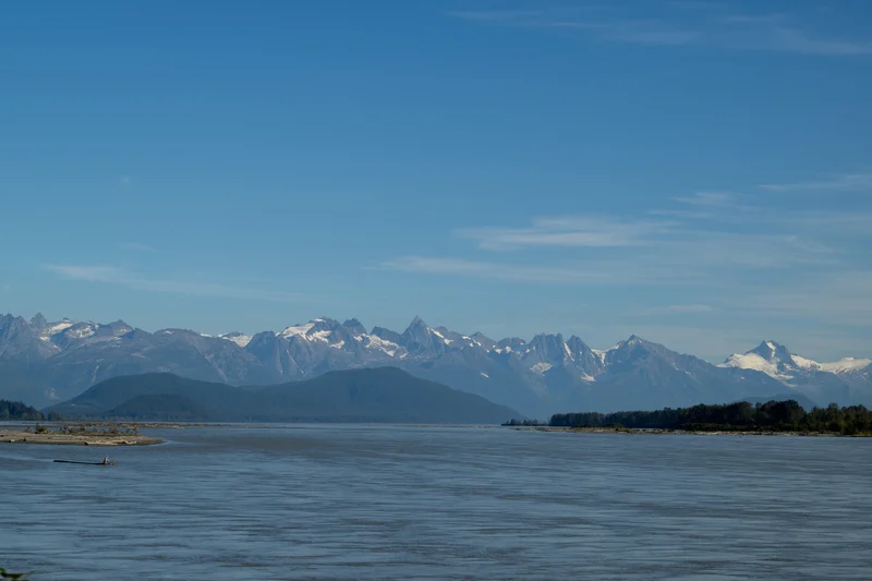 A wide landscape showing a calm body of water, forested hills, and a distant range of rugged, snow-capped mountains under a blue sky.