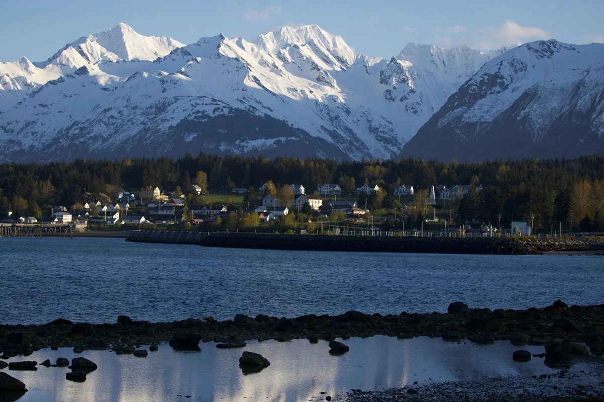 A scenic view of a coastal town with white houses and green trees, situated between a calm body of water and towering snow-capped mountains under a clear sky.