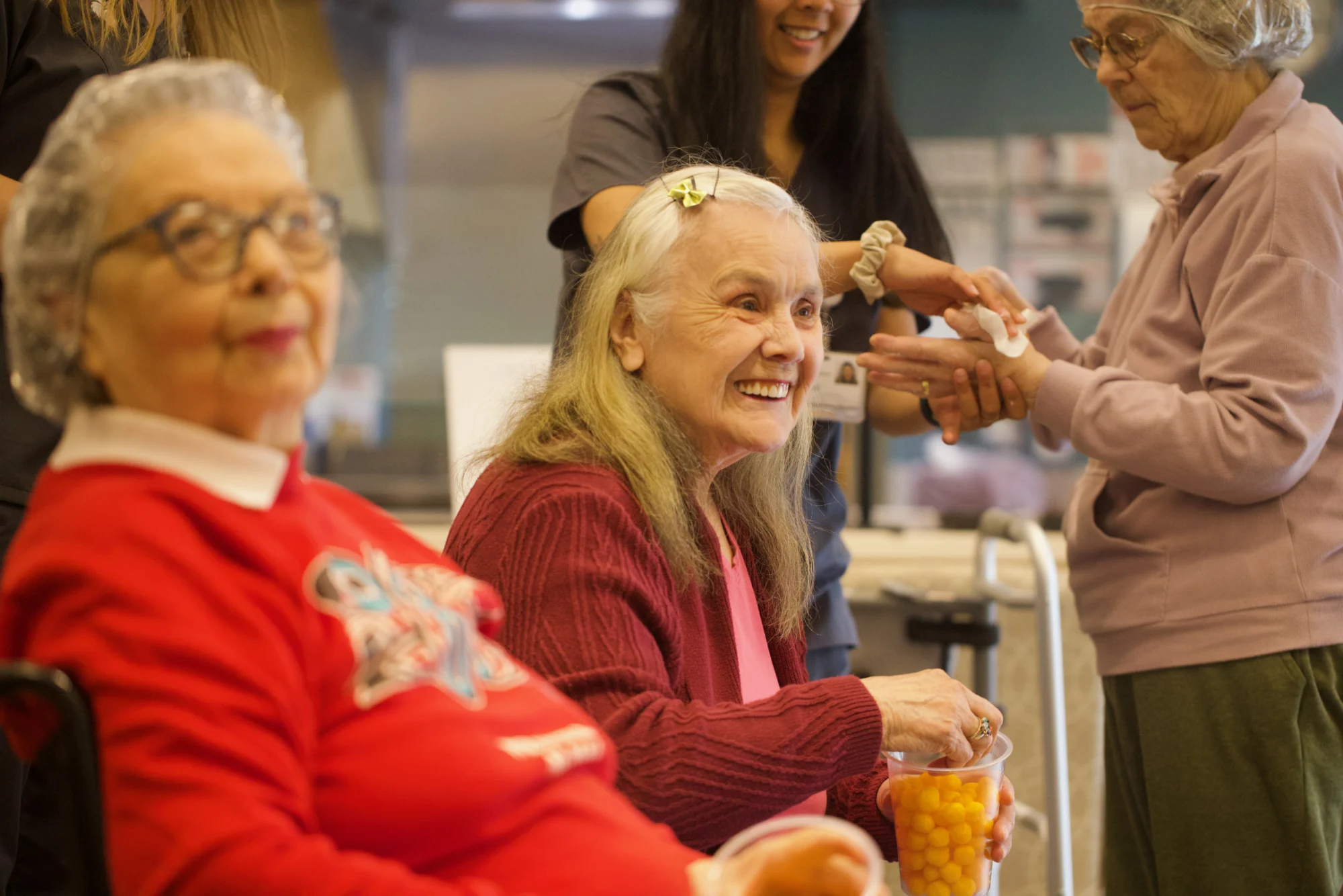 Three women, two elderly and one younger, interacting. An elderly woman in the center smiles broadly while holding a cup of snacks. A younger woman attends to the hands of another elderly woman on the right.