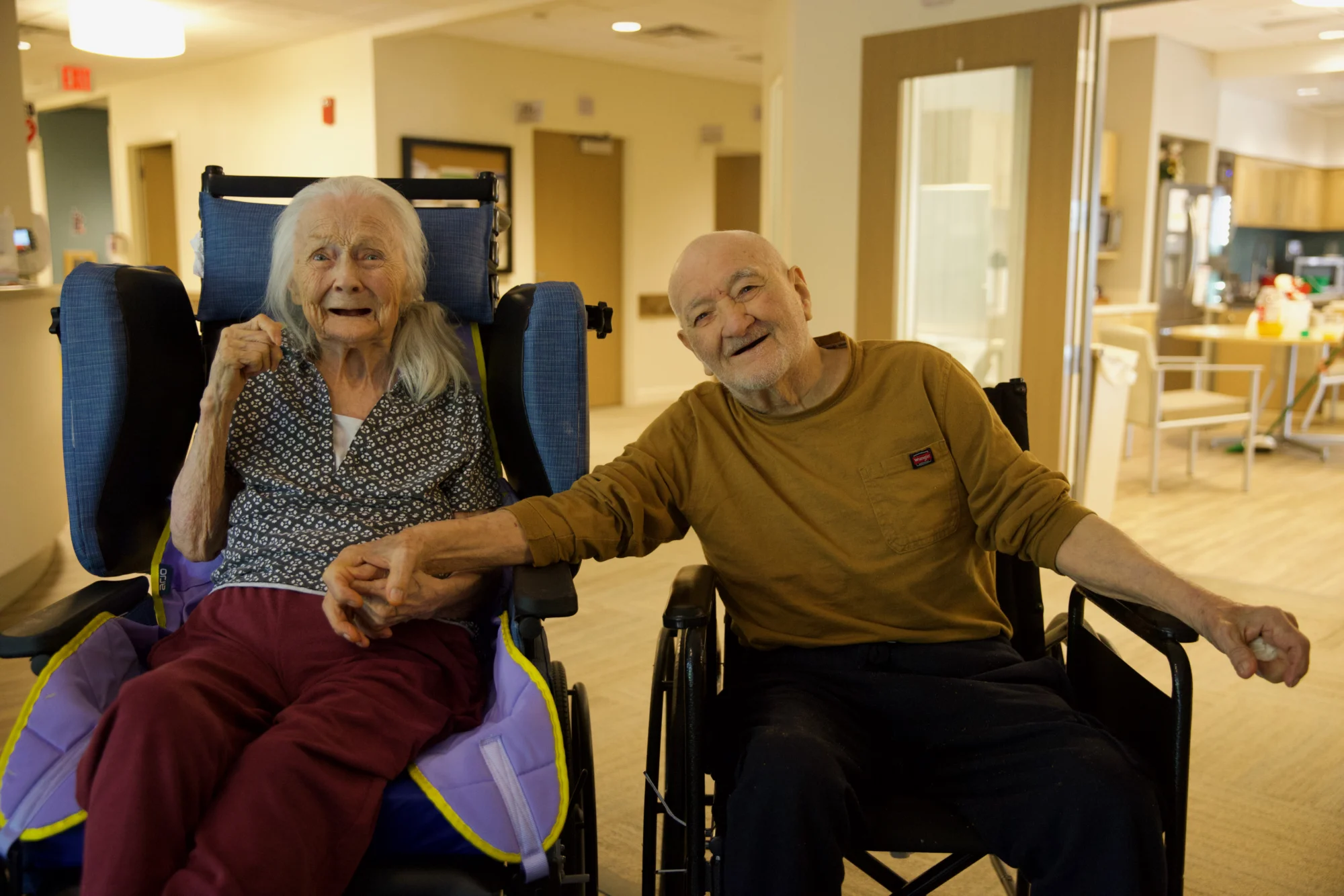 Two elderly individuals, a woman with white hair and a man with a white beard, sit side-by-side in wheelchairs, holding hands and smiling at the camera in an indoor facility.