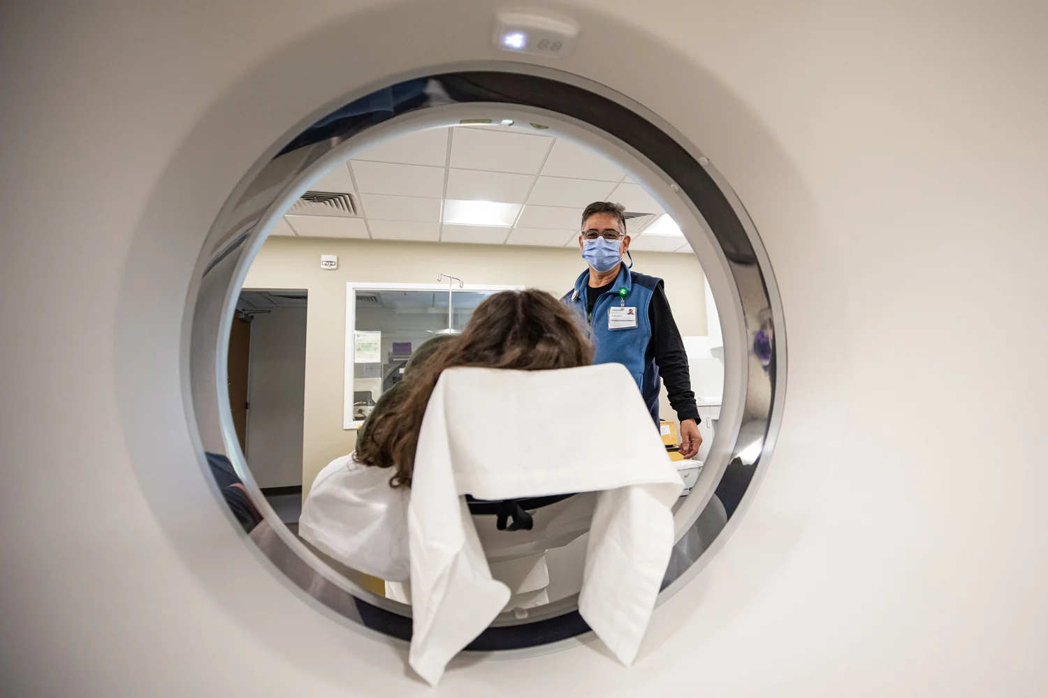A patient lies on a medical table, viewed through a CT scanner opening, with a masked healthcare professional standing behind them.