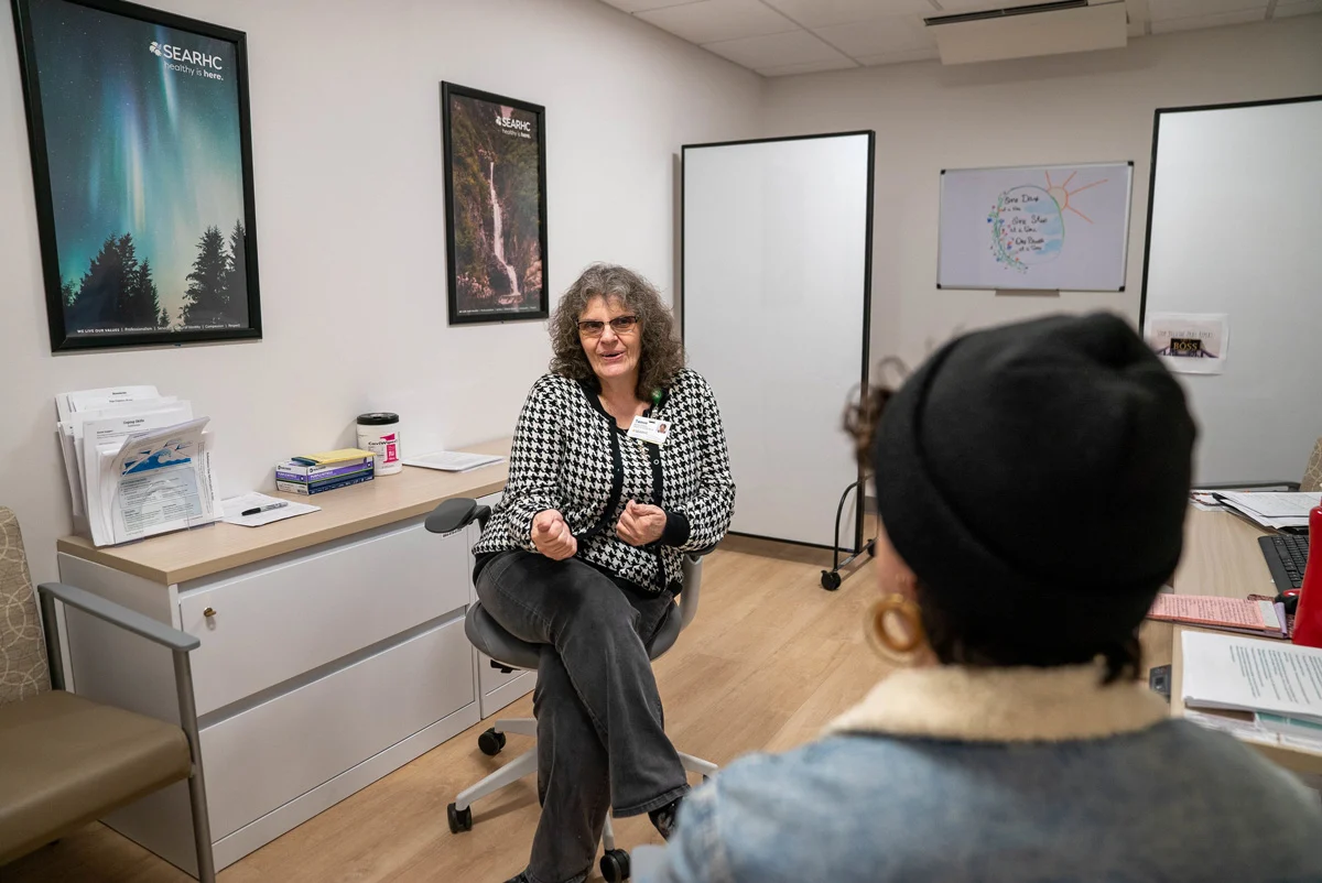 A woman with curly hair and glasses in a houndstooth cardigan sits in an office chair, facing a person seen from behind in a black beanie and denim jacket. They are in a room with framed nature posters and whiteboards.