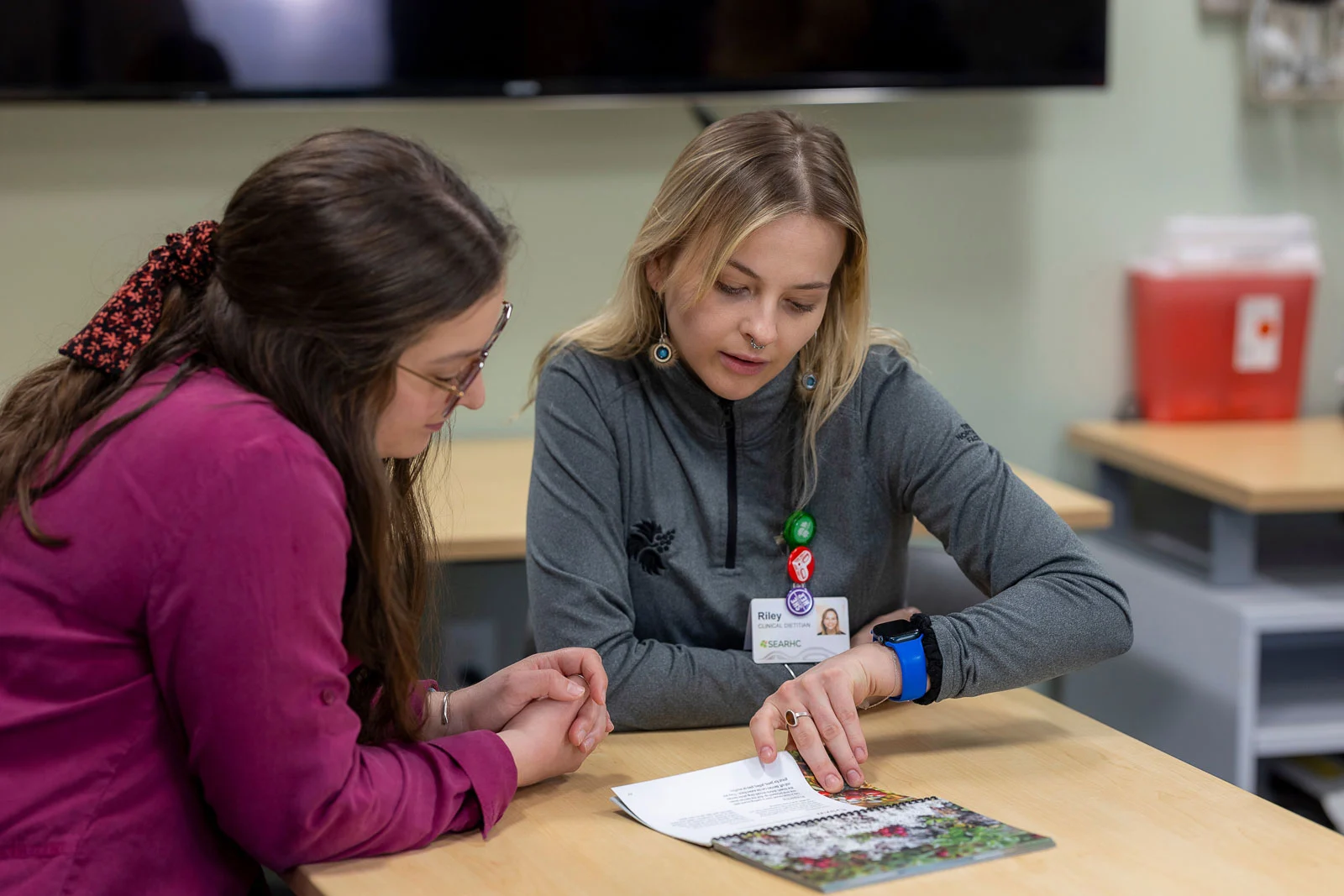 Two women at a light wooden table. One, a blonde woman in a grey shirt with an ID badge reading 'Riley, Clinical Dietitian,' points to an open booklet. The other, a woman with long dark hair in a magenta shirt, looks on.