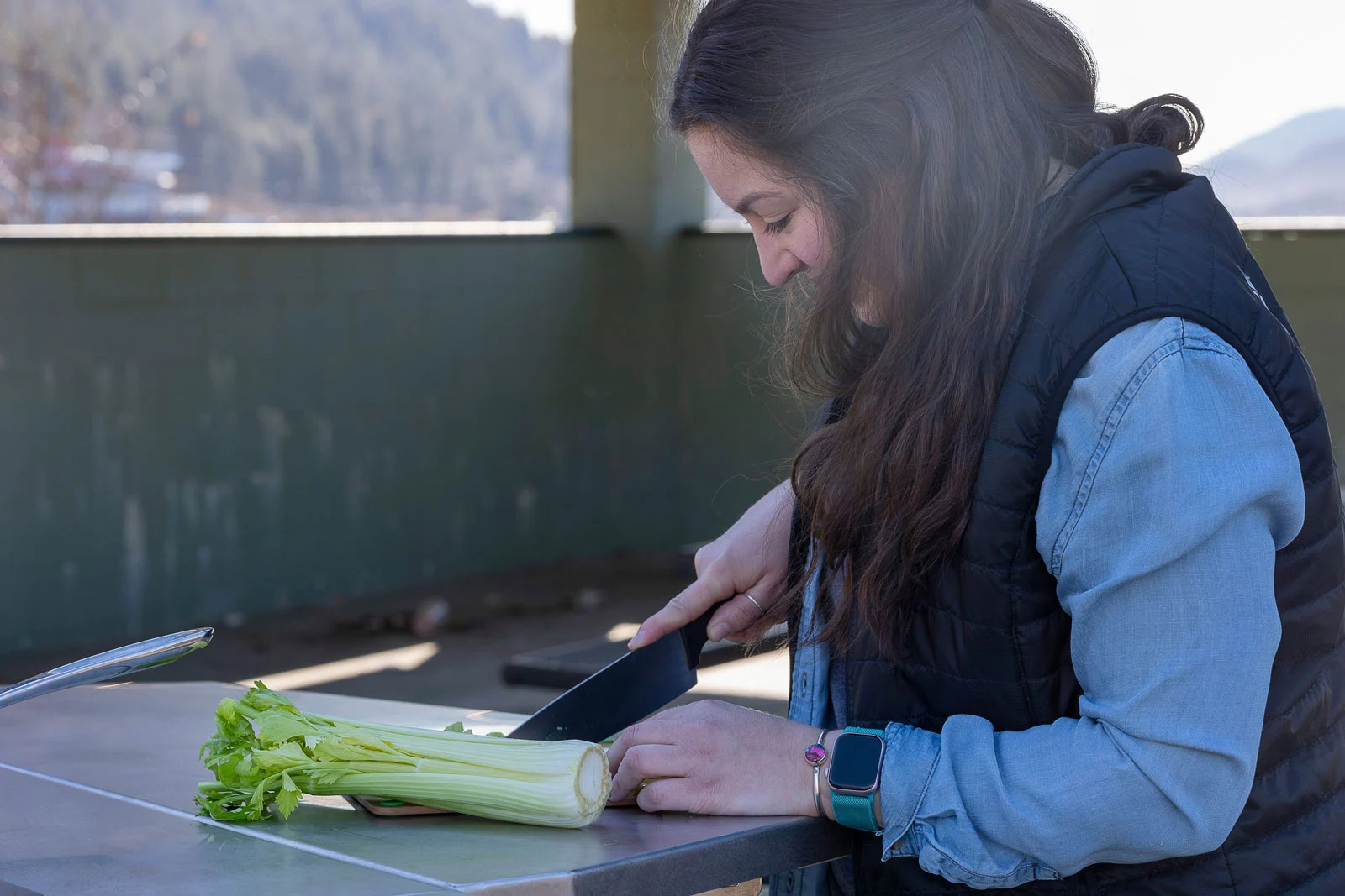 A woman cuts celery on an outdoor counter, wearing a vest and smartwatch, with mountains in the blurred background.