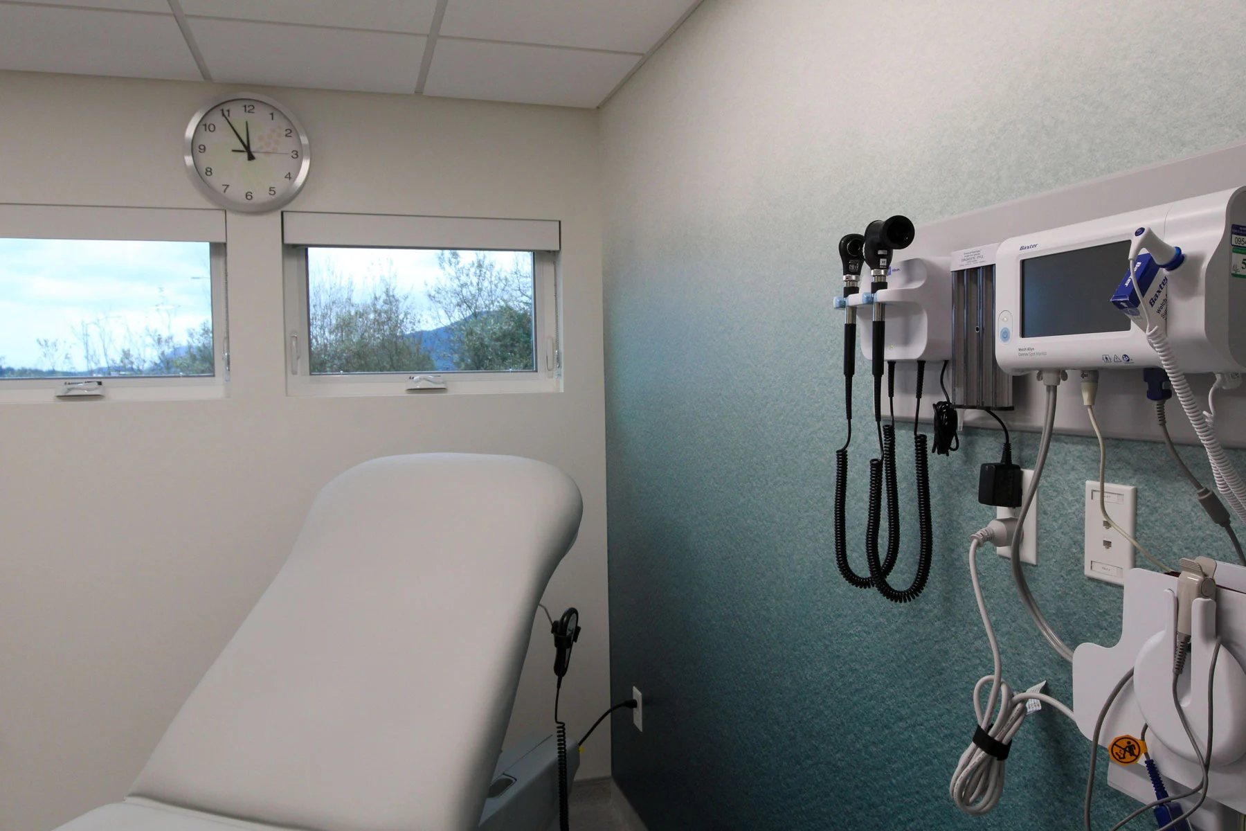 A medical examination room with a gray examination table, two windows, a wall clock, and wall-mounted diagnostic equipment including a vital signs monitor.