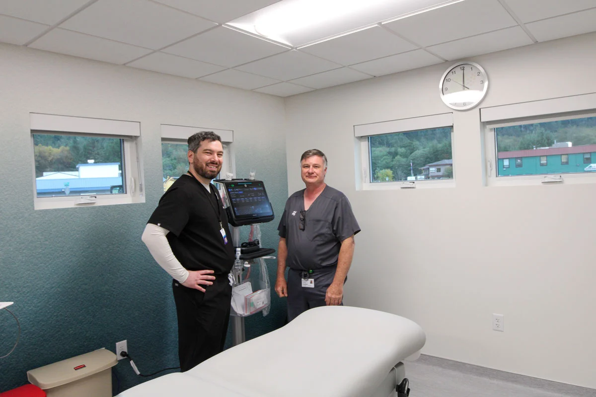 Two male medical professionals in scrubs stand in a clean, brightly lit examination room with a medical device and an examination table.
