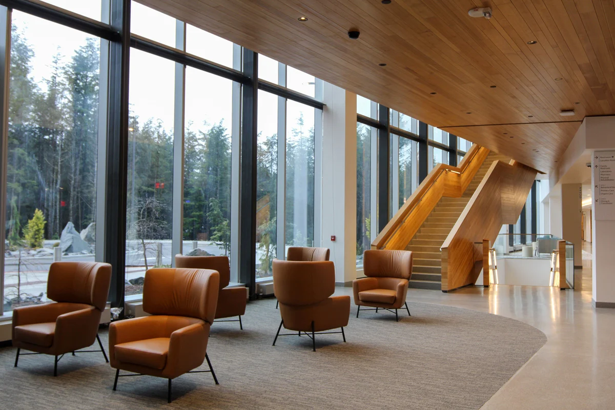 Modern lobby with large windows, brown leather armchairs, wooden staircase, and forest view.