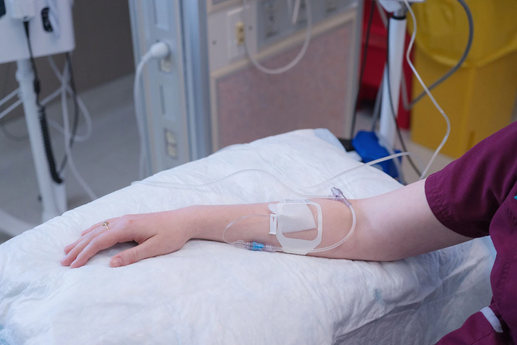 A person's arm with an IV line inserted, resting on a white medical bed. A ring is on the finger, and medical equipment is in the background.