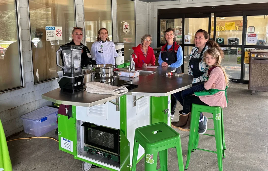 Six people, including a child, around a green and silver mobile cooking station with a blender and oven, under a covered outdoor area.