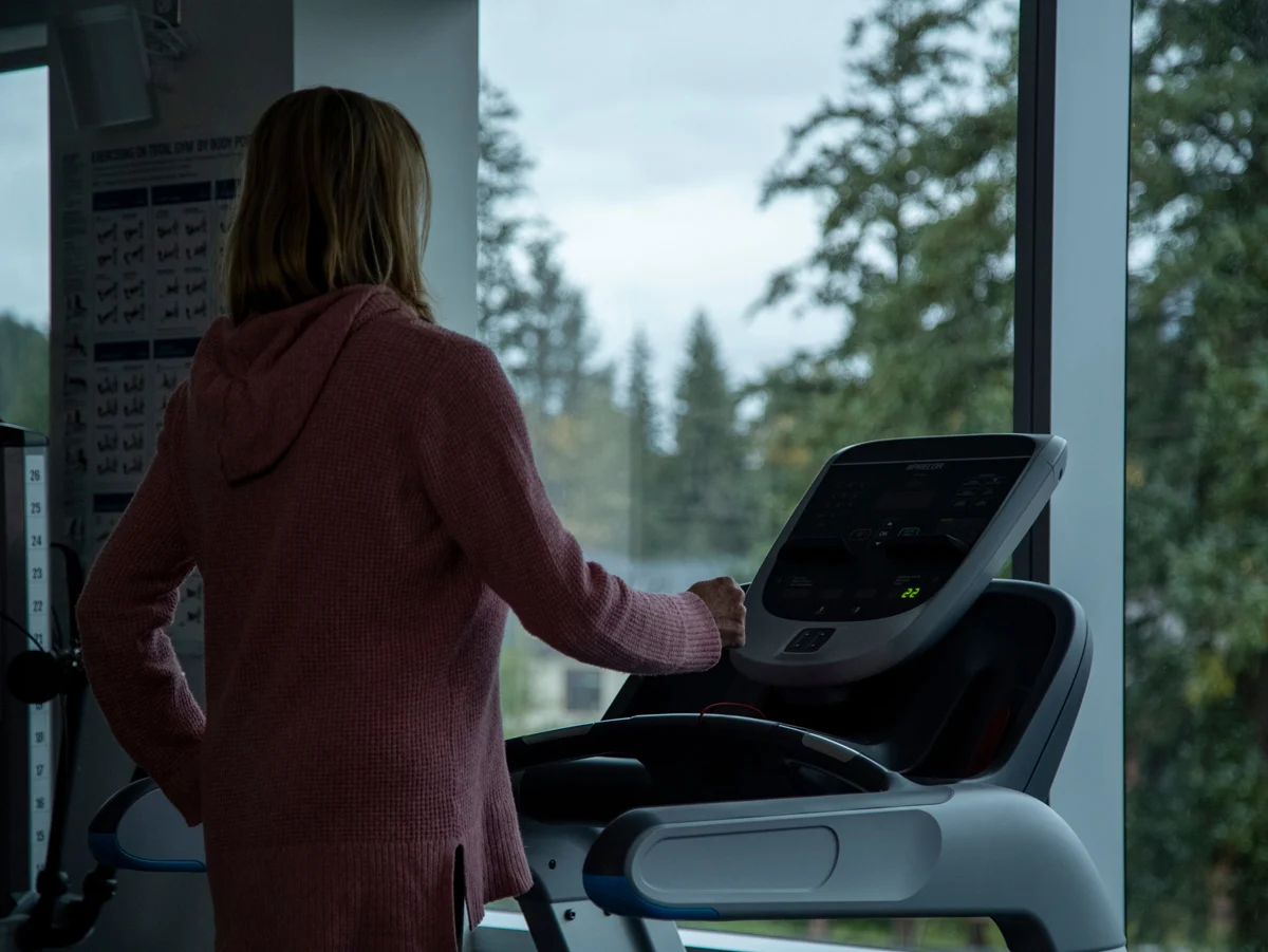 A person in a pink hoodie stands on a treadmill, looking out a large window at green trees under an overcast sky.