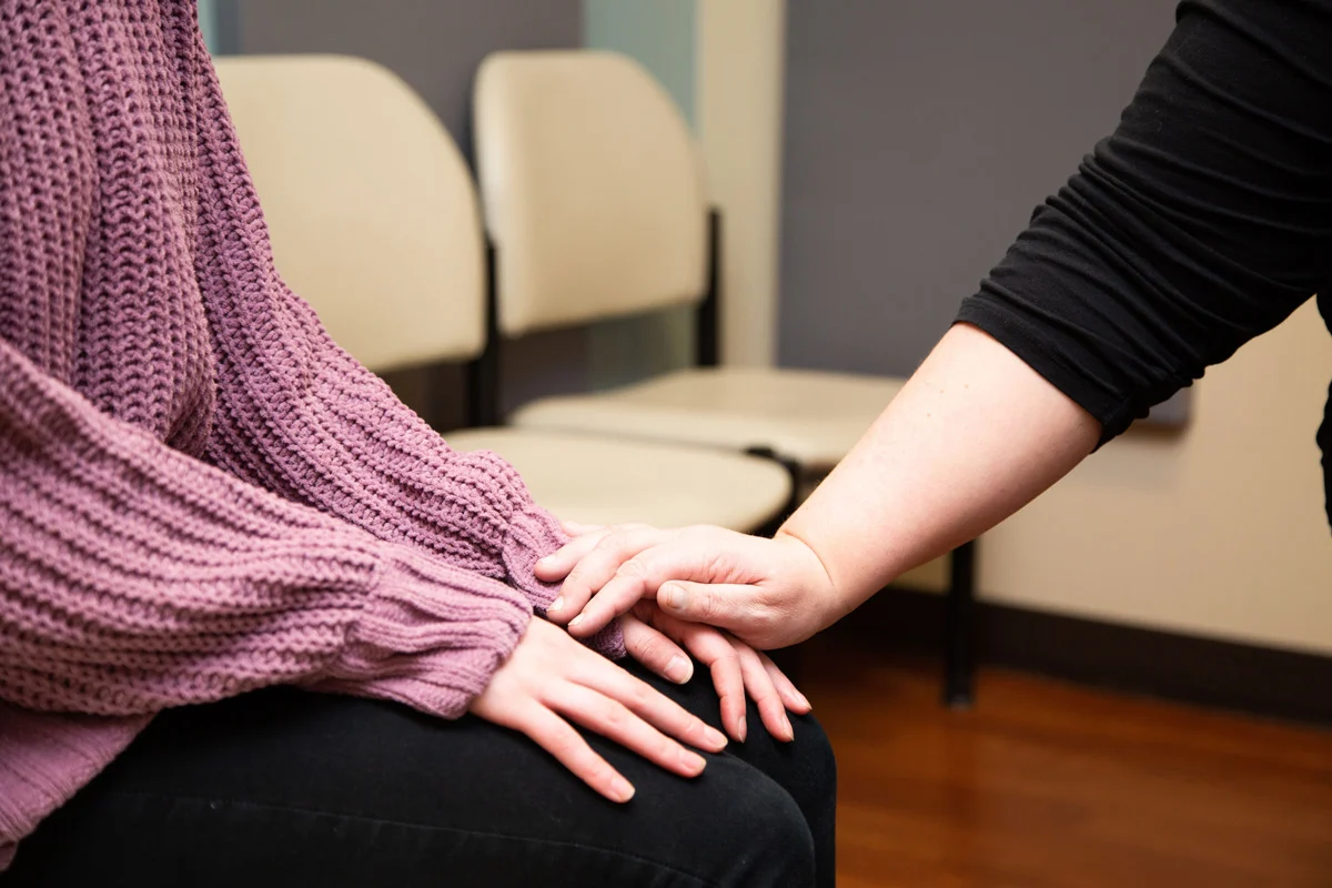 One person's hand gently rests on another person's clasped hands, offering comfort and support in a waiting room setting.