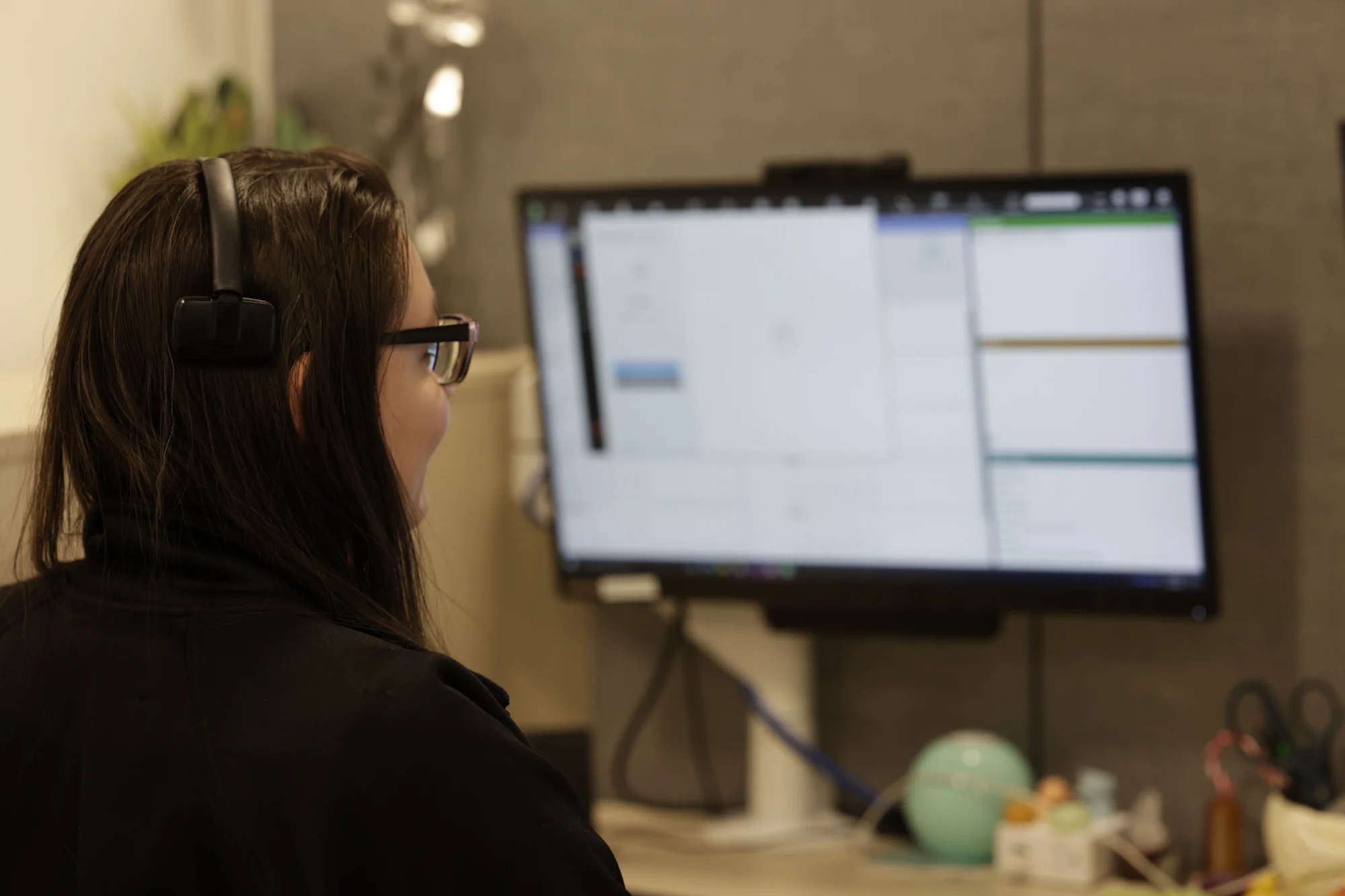A person with long dark hair, wearing black headphones and glasses, looking at a computer monitor displaying a software interface.