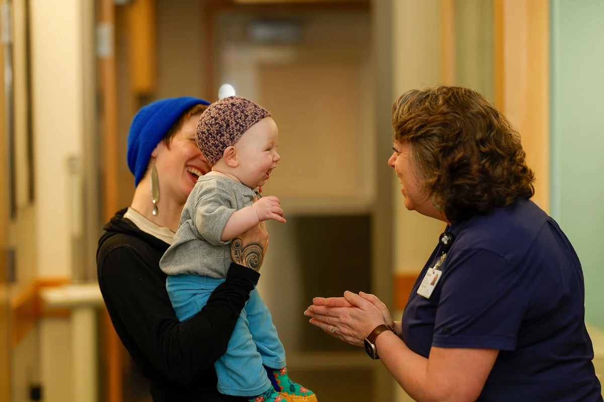 A person in a blue beanie holds a laughing baby wearing a crocheted hat, while a woman in blue scrubs smiles at them. All three are laughing in an indoor setting.