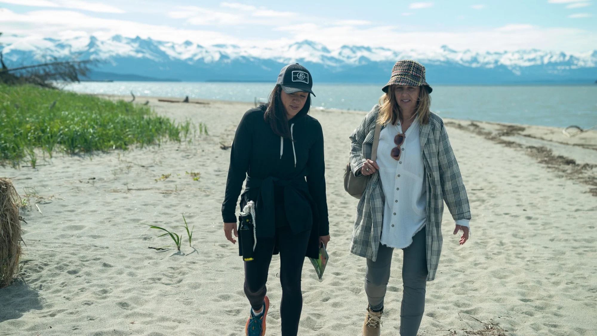 Two women walk on a sandy beach with a large body of water and snow-capped mountains in the background under a partly cloudy sky.