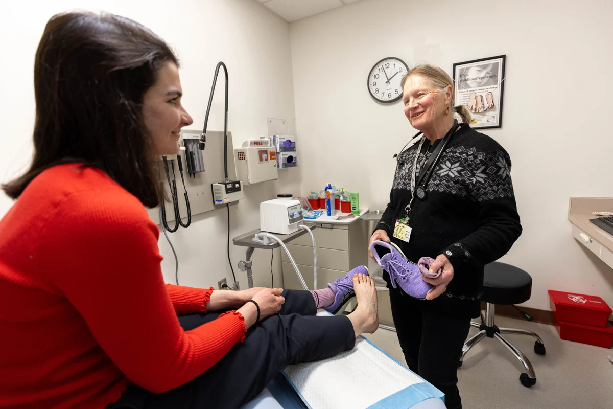 A medical professional holding purple shoes smiles at a patient sitting on an exam table, who is wearing one purple shoe and smiling back.