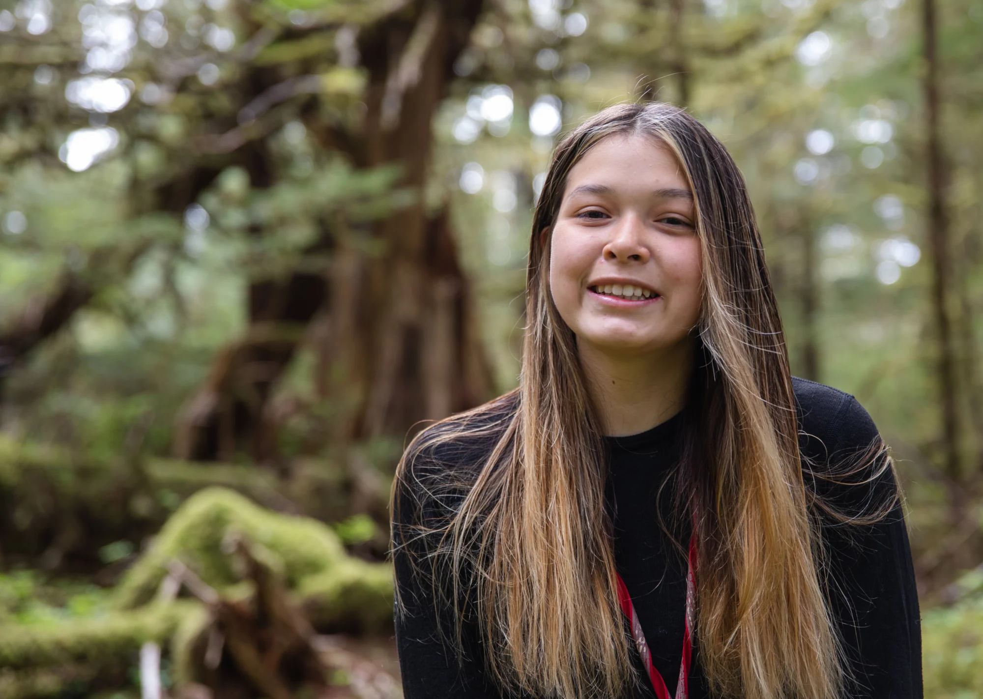 A young person with long, highlighted hair smiles at the camera in a blurred forest setting.
