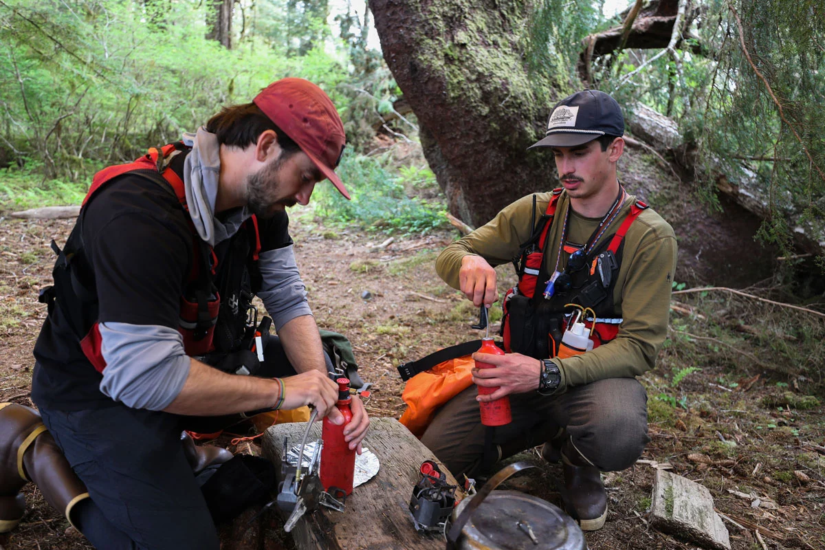 Two men in outdoor gear crouch in a forest, operating portable camping stoves with red fuel bottles.