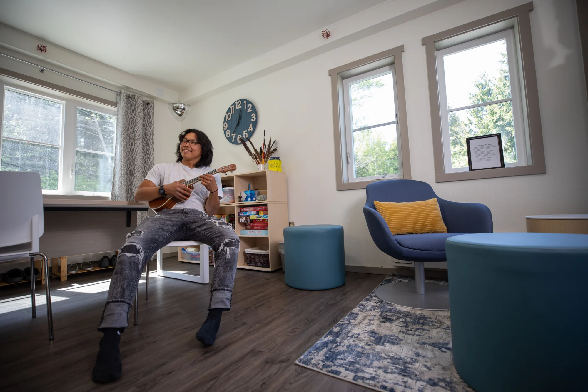 A young man with long hair and glasses plays a ukulele while sitting in a bright room with windows, a blue clock, and various furniture.