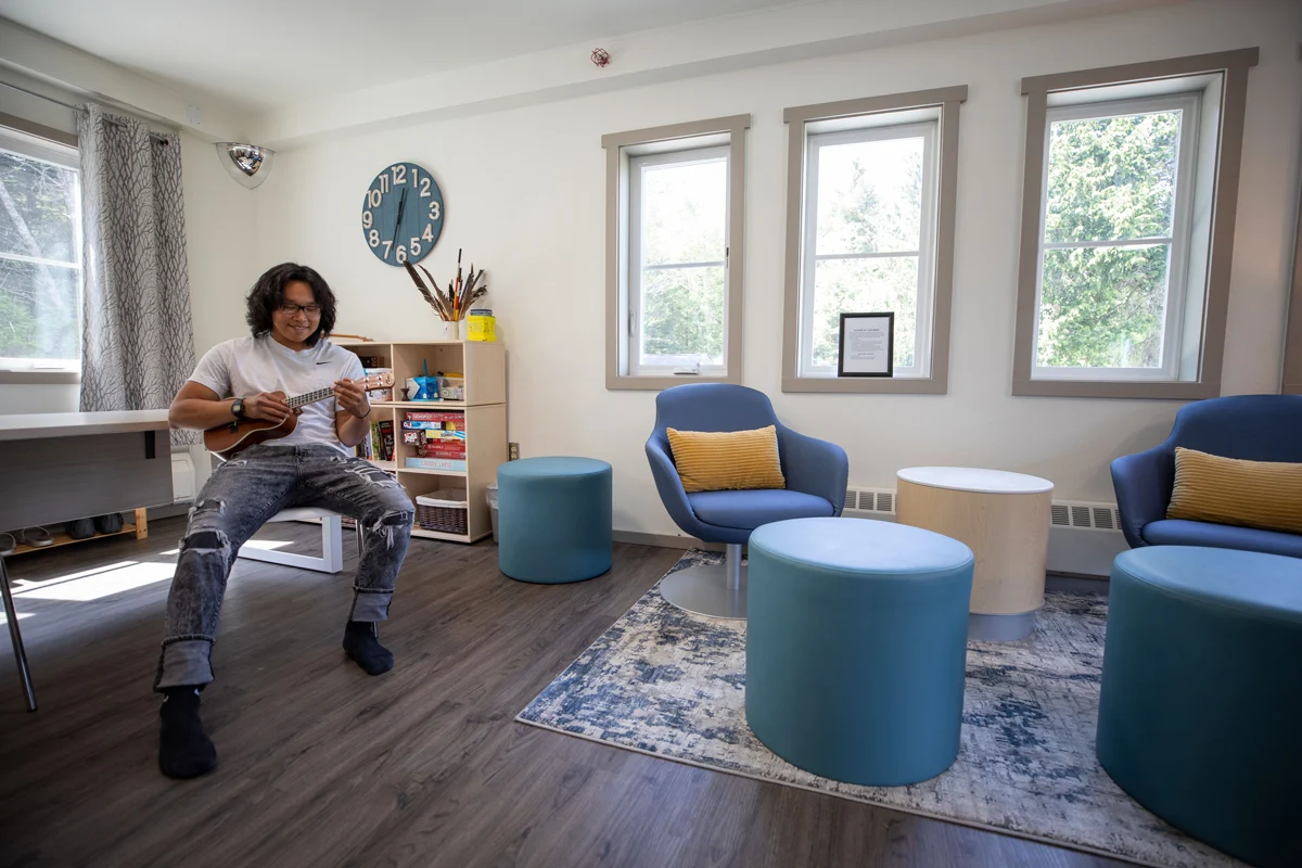 A young man with glasses smiles while playing a ukulele in a brightly lit room with blue armchairs and large windows.