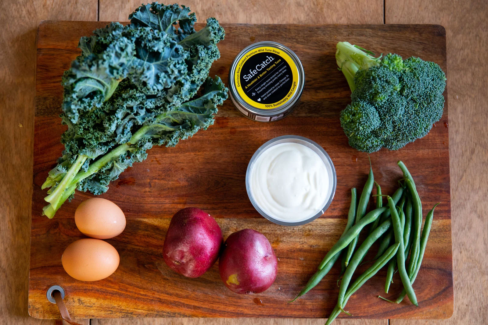 Overhead view of fresh ingredients on a wooden cutting board, including kale, broccoli, green beans, red potatoes, eggs, a bowl of white cream, and a can of SafeCatch tuna.