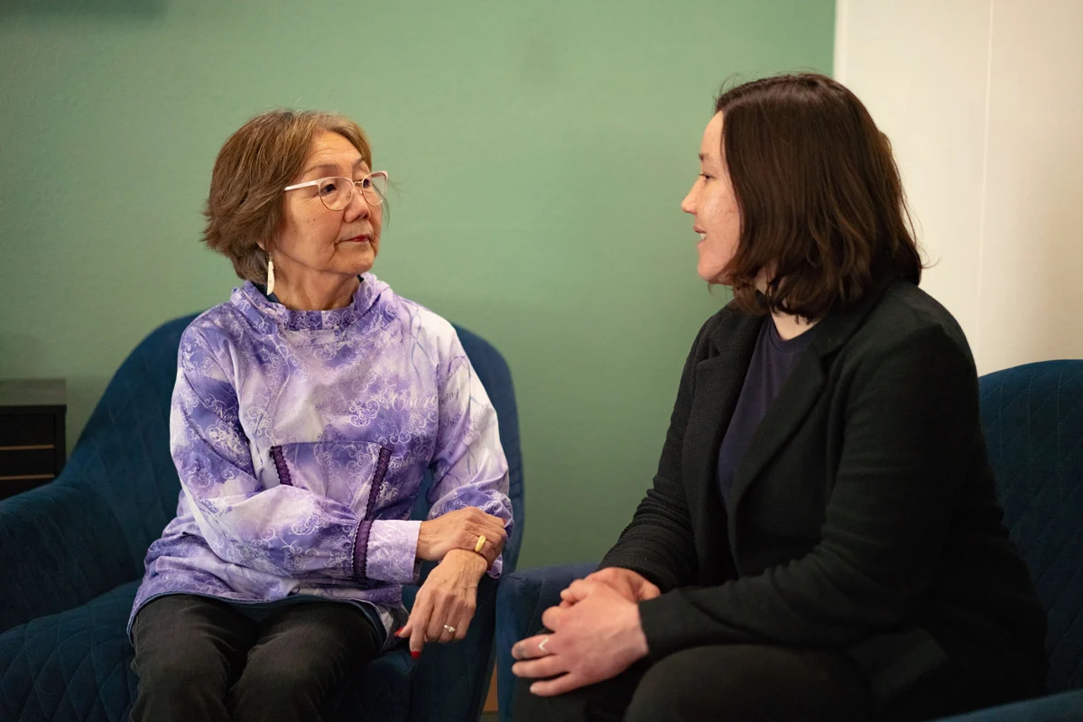 Two women, one older with a purple patterned top and one younger in a dark blazer, sit facing each other and talking.