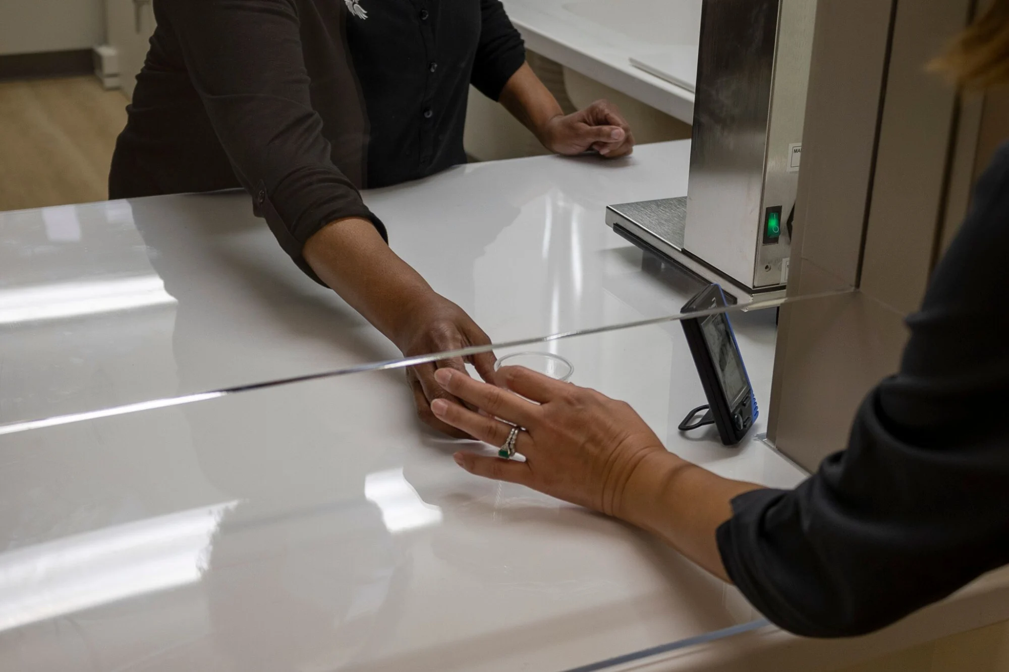 Two people's hands reaching across a white counter with a clear barrier; one hand wears a ring with a green stone, and a small clear cup is between them.