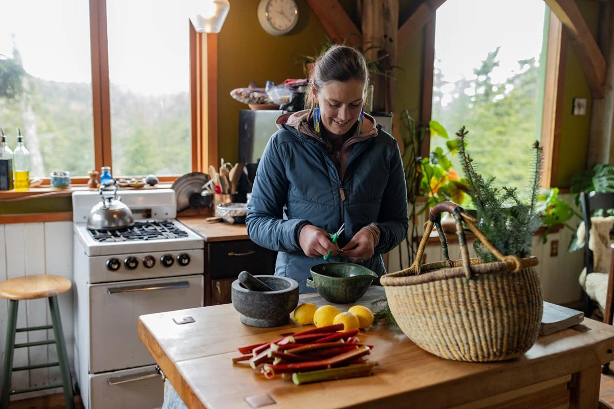 A woman in a blue jacket trims ingredients into a bowl in a rustic kitchen with rhubarb, lemons, and a mortar and pestle on a wooden table.