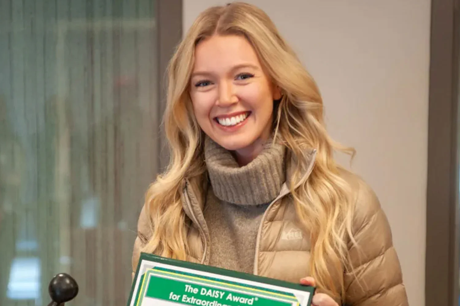 A smiling young woman with long blonde hair, wearing a tan puffer jacket and turtleneck, holds a green and white DAISY Award certificate.