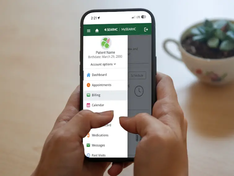 Hands holding a smartphone displaying the MySEARHC healthcare app with a menu showing patient information, appointments, and billing options.