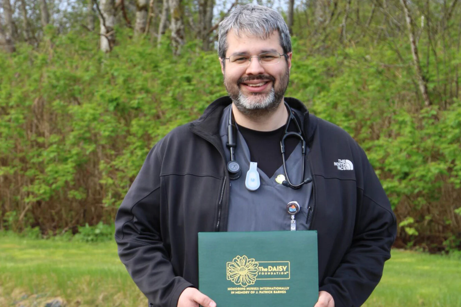 A smiling man, a healthcare professional, holds a green DAISY Foundation award certificate outdoors.