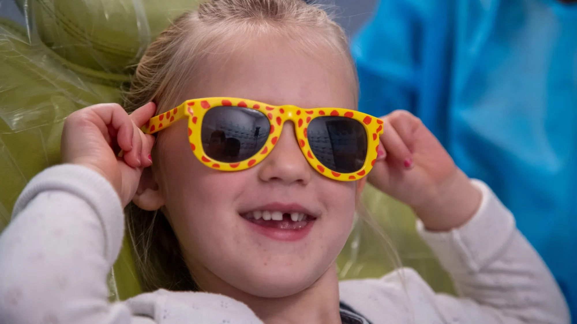 A smiling young girl with blonde hair wears yellow polka dot sunglasses, revealing missing front teeth.