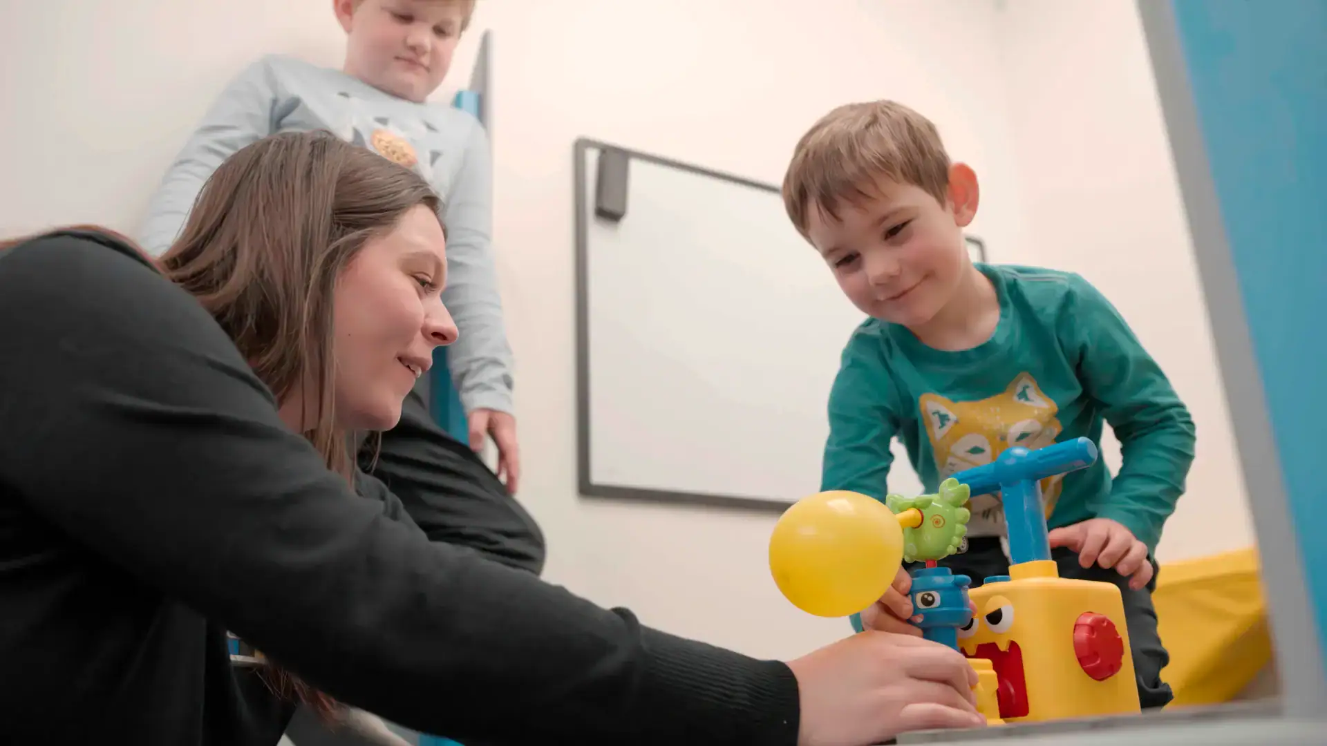 A woman and two young boys playing with a yellow and blue balloon pump toy indoors.