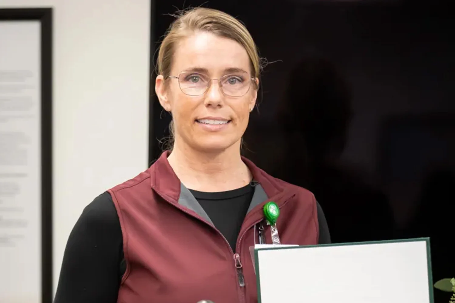 A smiling woman with blonde hair and glasses, wearing a maroon vest, holds a document with a green border.