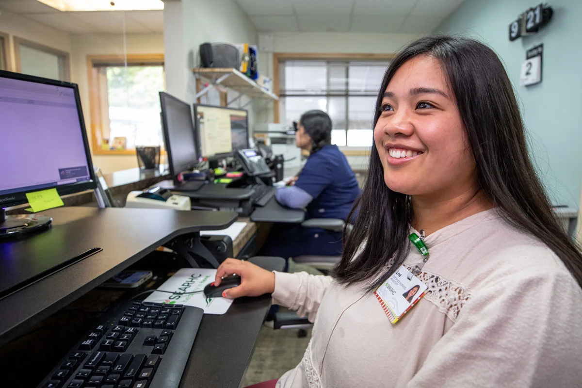 A smiling young woman, Lea, a Certified Nursing Assistant, works at a computer desk in an office.