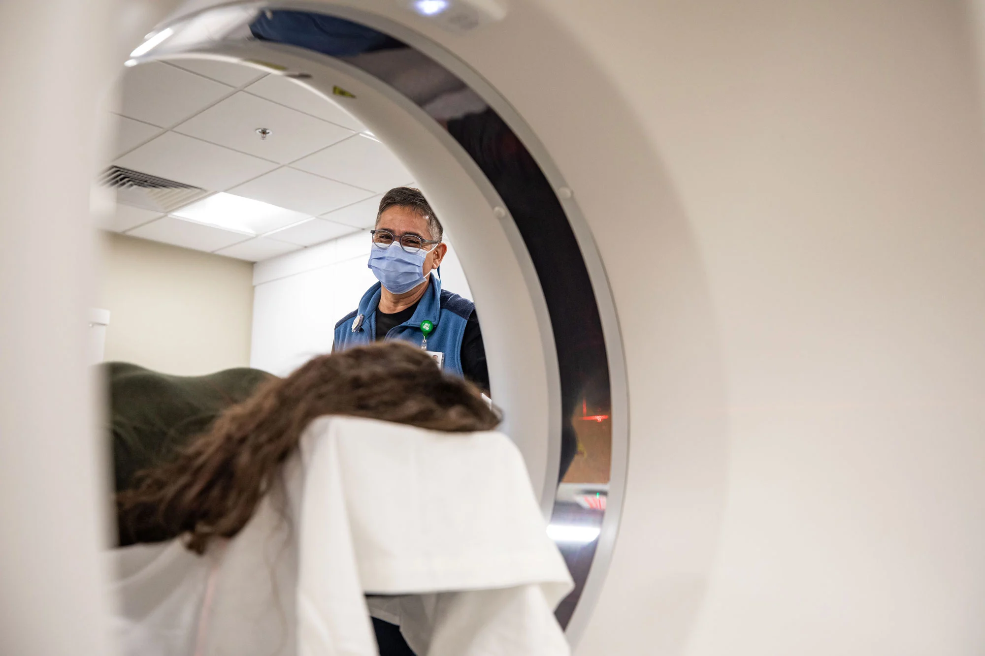 A medical professional wearing a blue mask and glasses stands behind a patient on a scanner table, viewed through the circular opening of a white medical imaging machine.