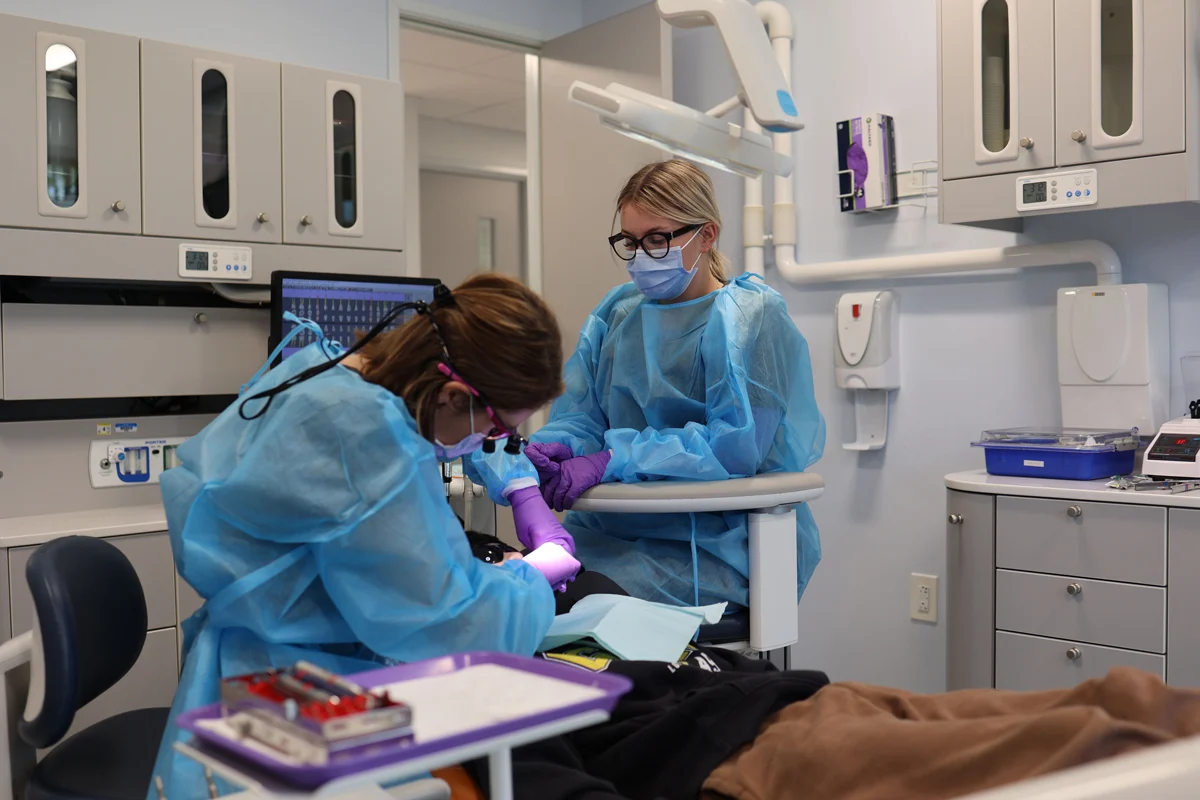 Two dental professionals in blue gowns and masks, one performing a procedure on a patient, the other observing in a dental clinic.