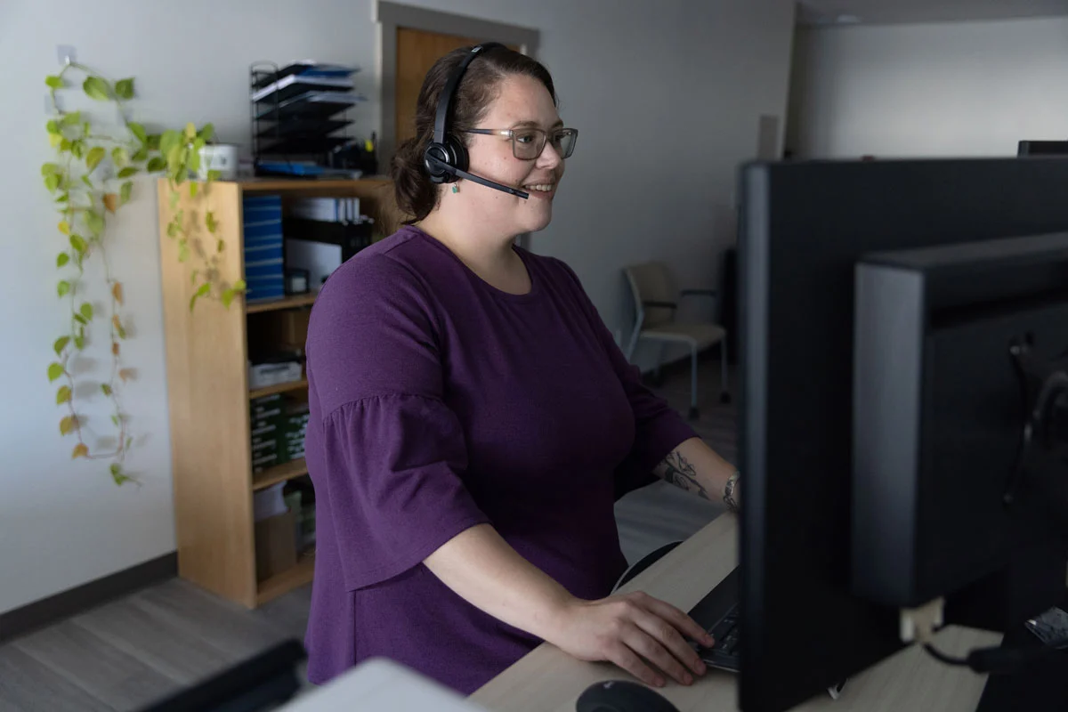 A woman with a headset and glasses sits at a desk, looking at a computer monitor and typing.