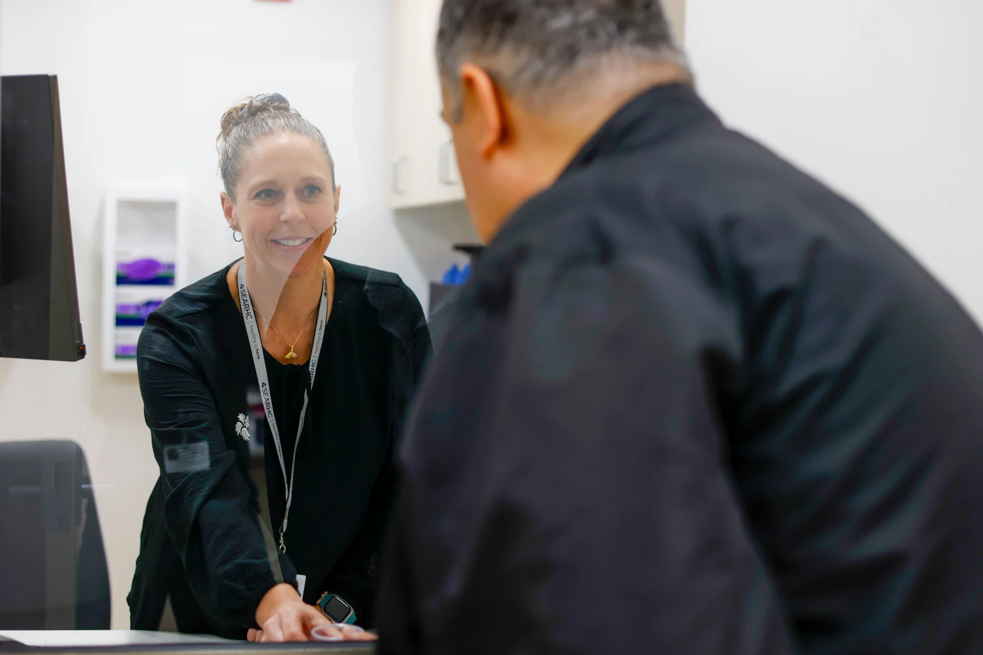 A smiling woman with a lanyard talks to a man across a counter, separated by a clear barrier.