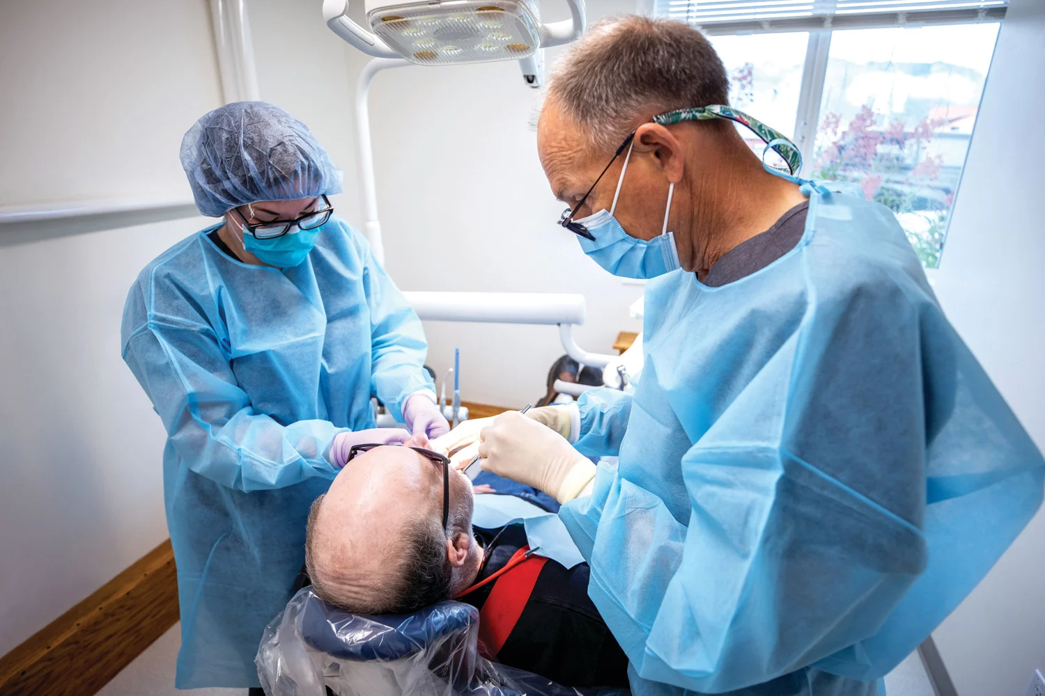 Two dental professionals in blue gowns, masks, and gloves work on a patient lying in a dental chair.